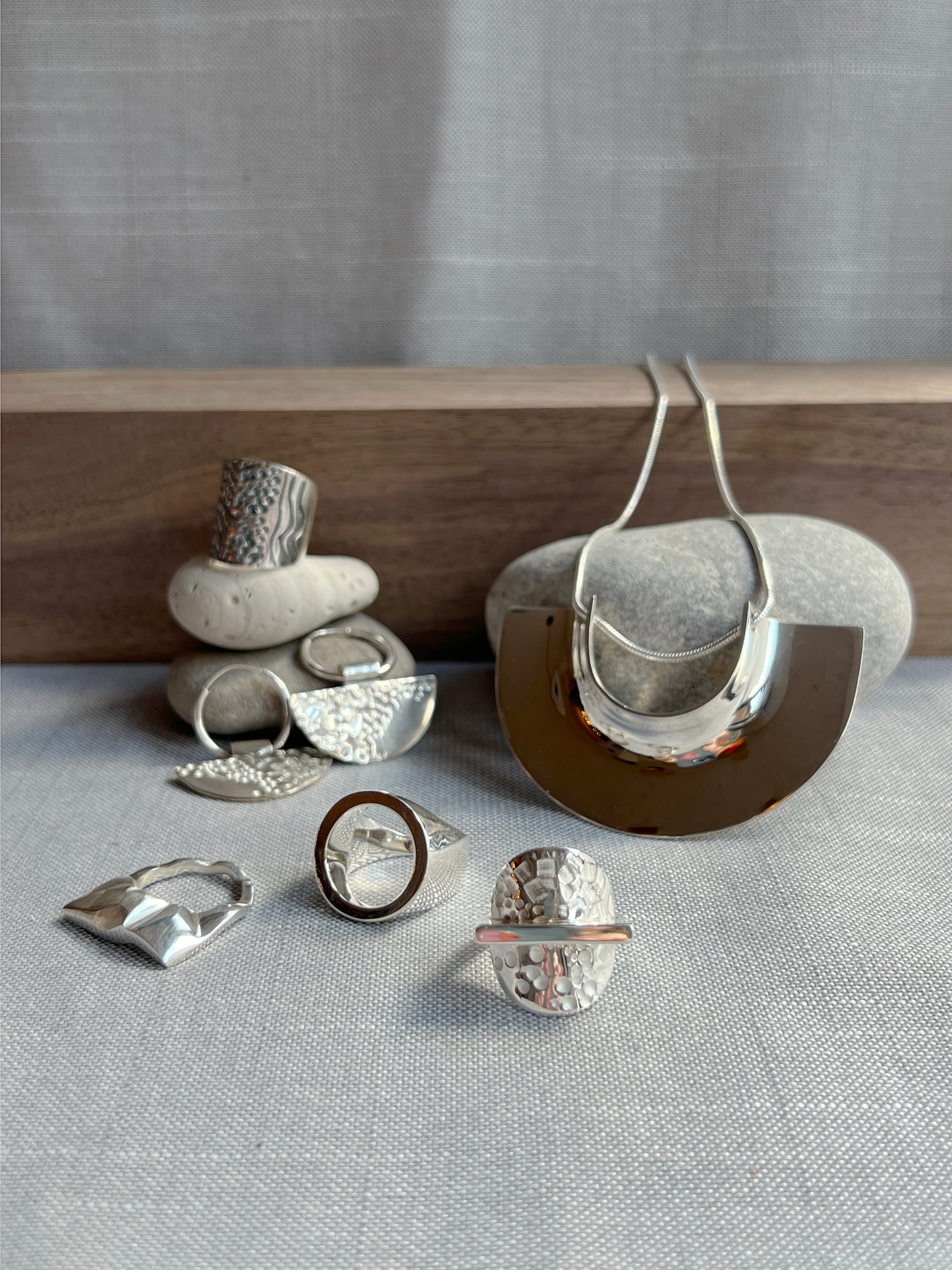 3 rings in the left foreground laying flat with 2 dangle earrings propped against 2 grey small rocks, with 1 ring on top of the grey rocks. on the right side, pendant necklace is propped up against 1 grey rock