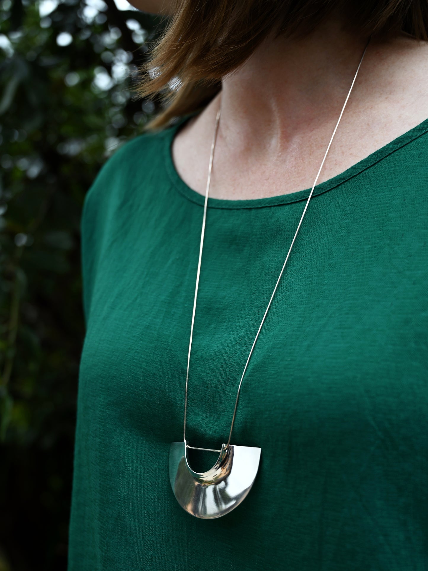 Full view of assemblage pendant around model's neck and green blouse worn with blurred greenery in background