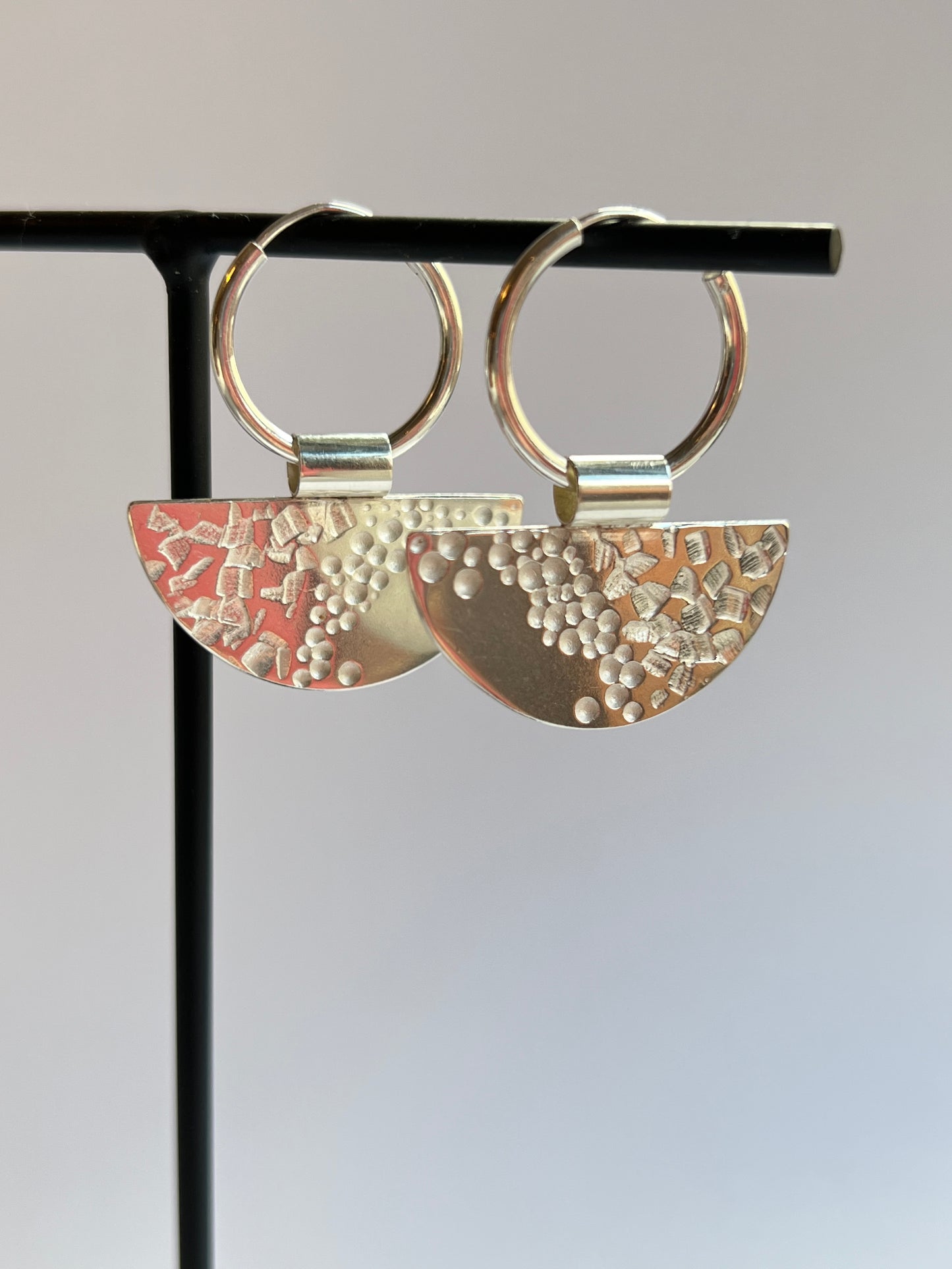 half moon disc hoops hanging up on a thin black stand, facing to the right, both earrings showing the front design