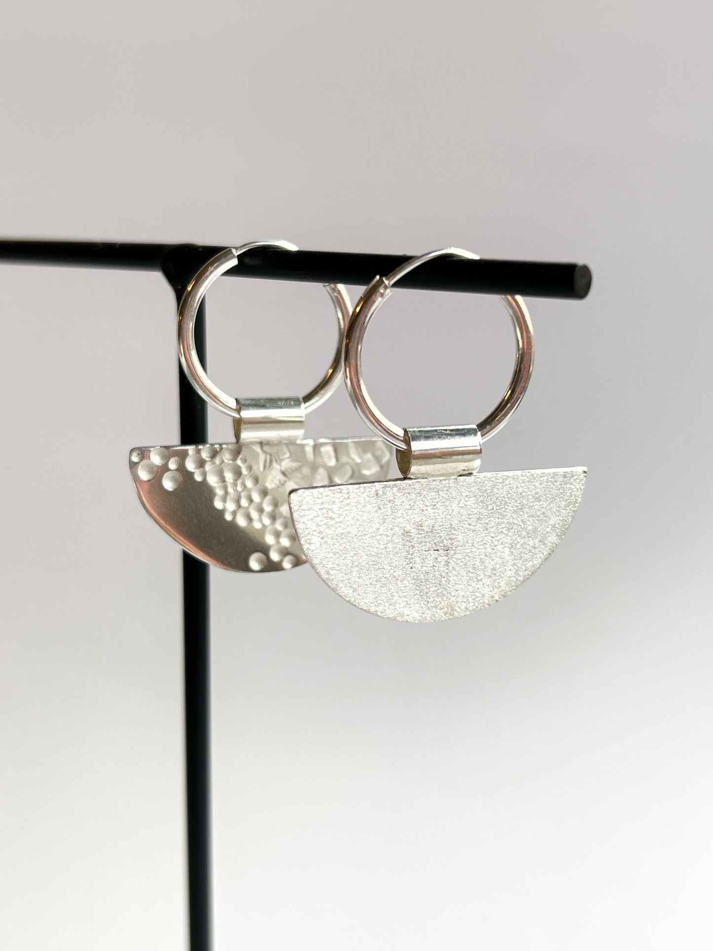 half moon disc hoops hanging up on a thin black stand, facing to the right, one earring showing the back and the other the front designs