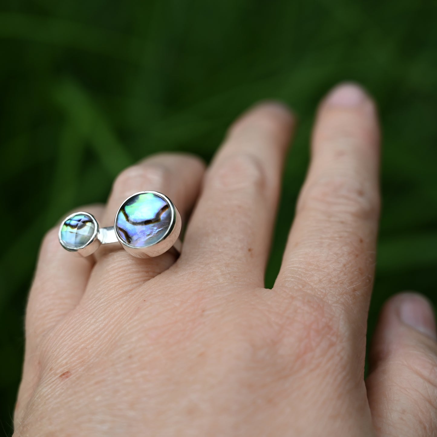 Component ring with double abalone stones on hand, side view with greenery in background.
