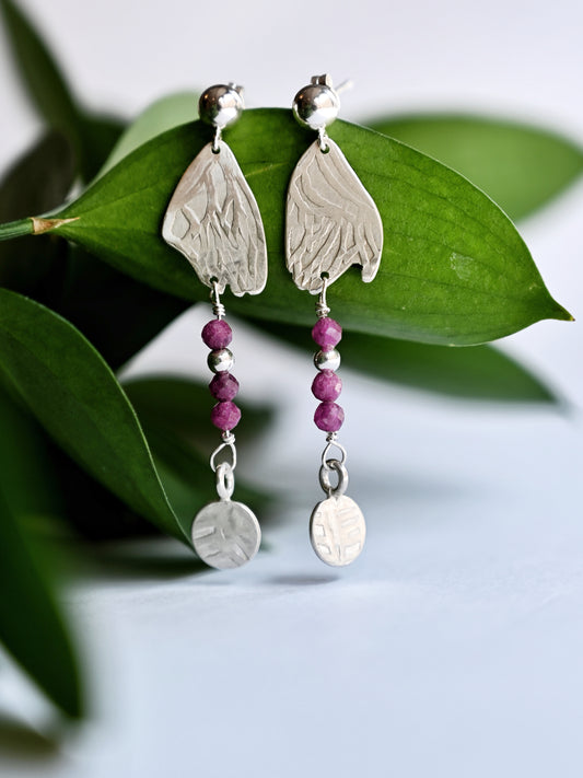 Silver dangling earrings with ruby hanging from greenery with white background