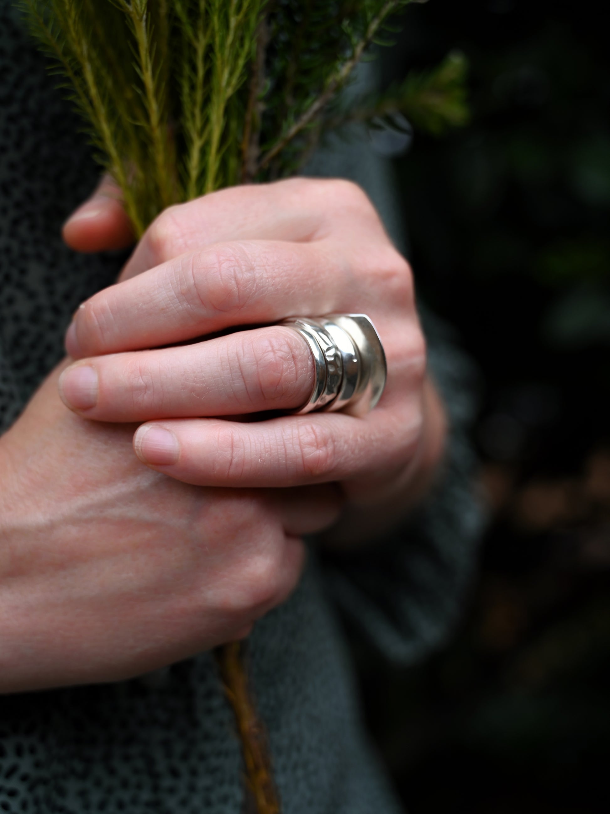 Element 1 ring in silver worn on ring finger together with other element rings, holding greenery and green and black dots blouse in background