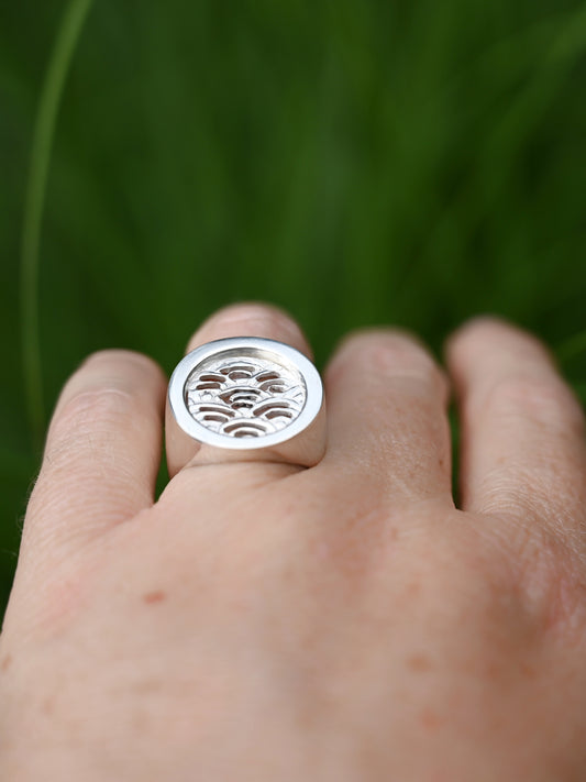 Silver node ring with cutout wave insert from top on hand with greenery in background