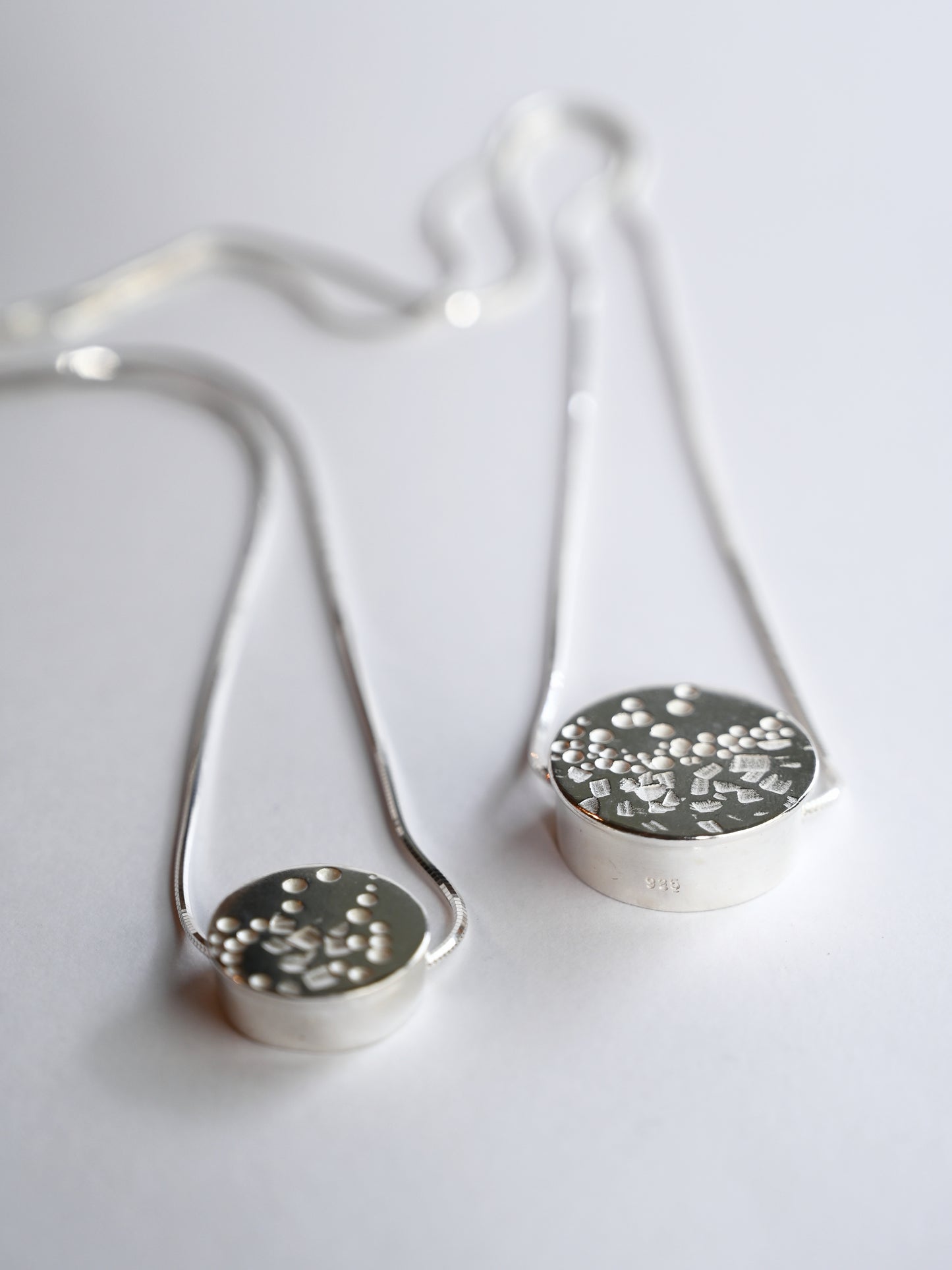 two silver circle necklaces laying flat on a white background, the back of the necklace chain is blurred