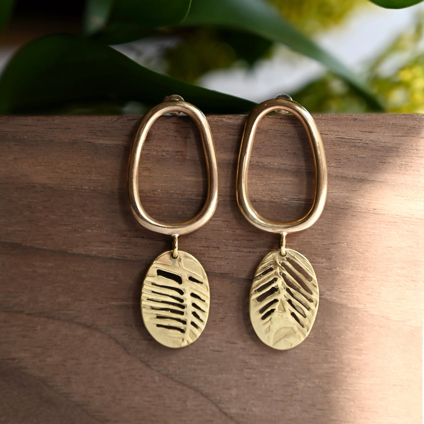 Bronze oval drop earrings with cutout leaf discs from front on wood block and greenery in background